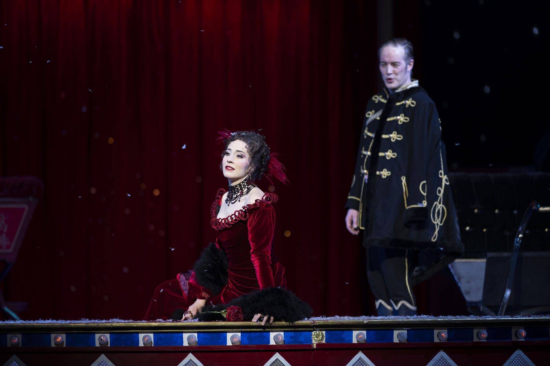 A woman in a red dress sits at the edge of a circus ring and gazes into the distance. Behind her stands a man in an elegant black outfit, looking at her.