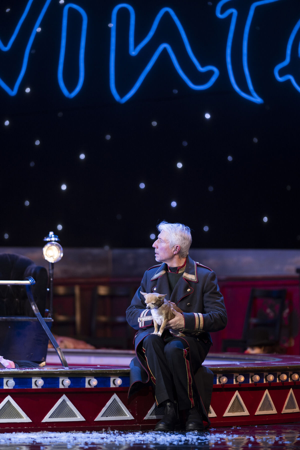 A man in a green uniform is sitting in a circus ring. He is holding a light brown Chihuahua on his lap.