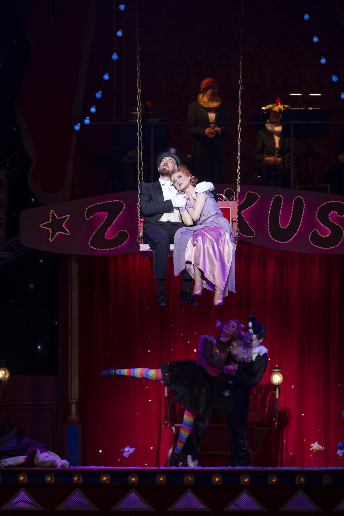 A man in a black suit and black top hat is sitting on a swing. Next to him is a woman in a pink and purple dress leaning against him. Two clowns are dancing beneath them.