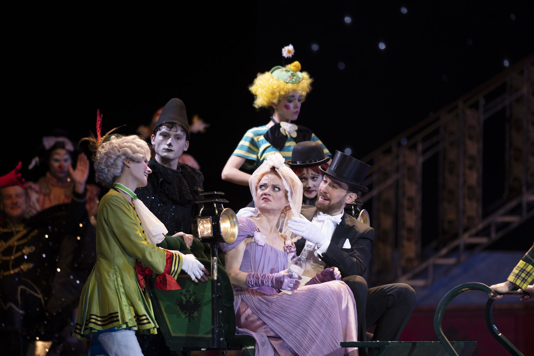 A man in a black suit and a woman in a purple dress with a veil are sitting in a carriage. The woman looks confused at a clown sitting next to her.