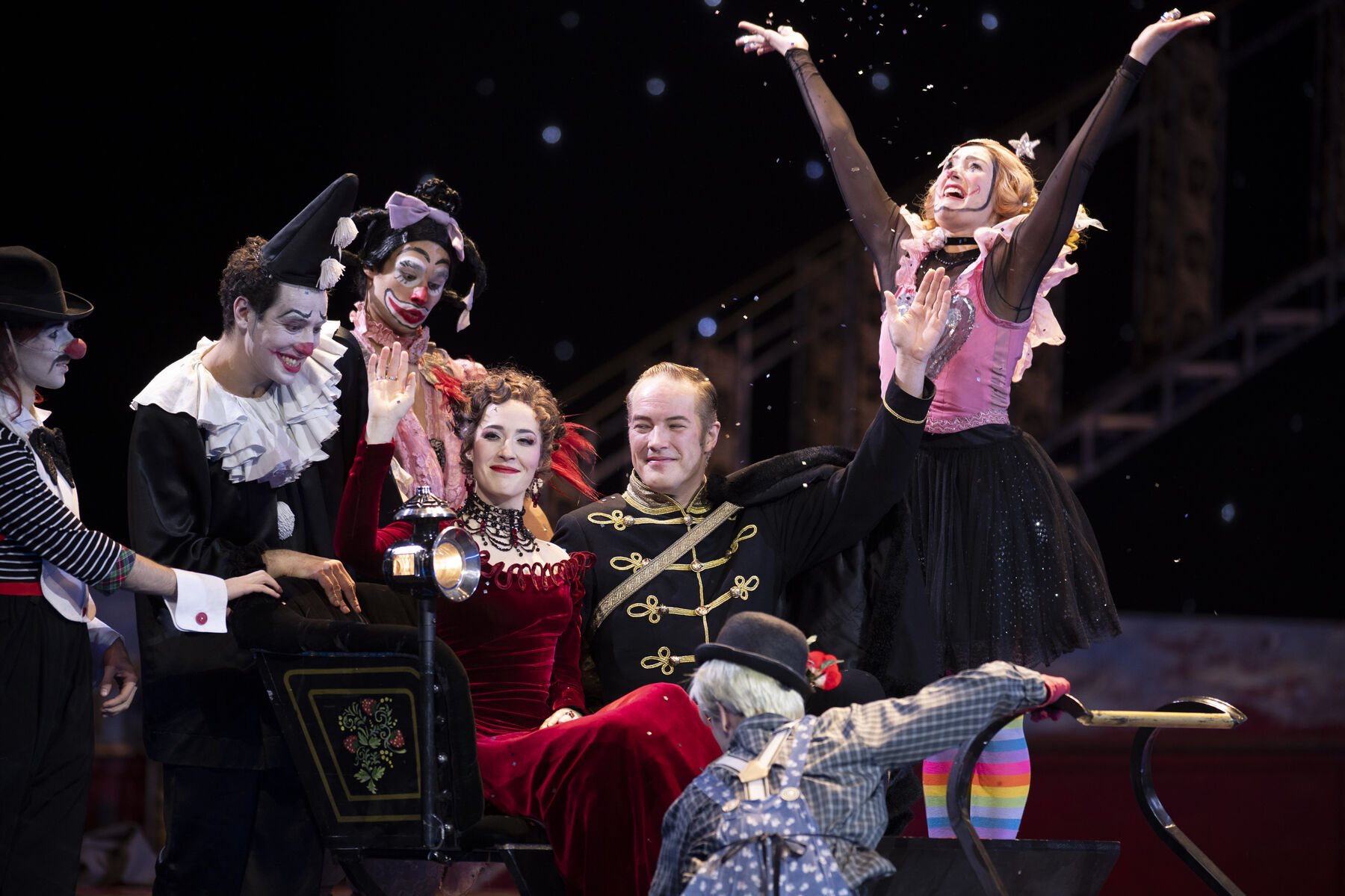 A woman in a red evening gown and a man in a black uniform sit waving in a carriage. Joyful clowns surround them.