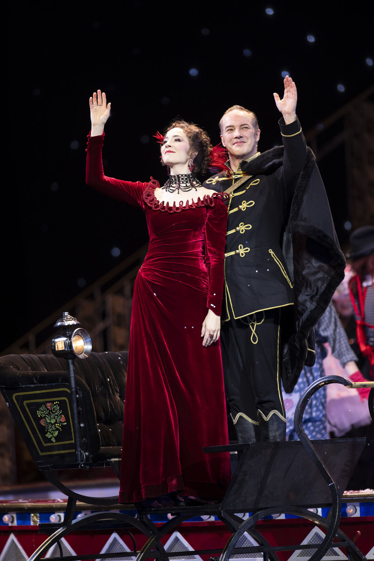 A woman in a red evening gown and a man in a black uniform are standing on a carriage, waving.