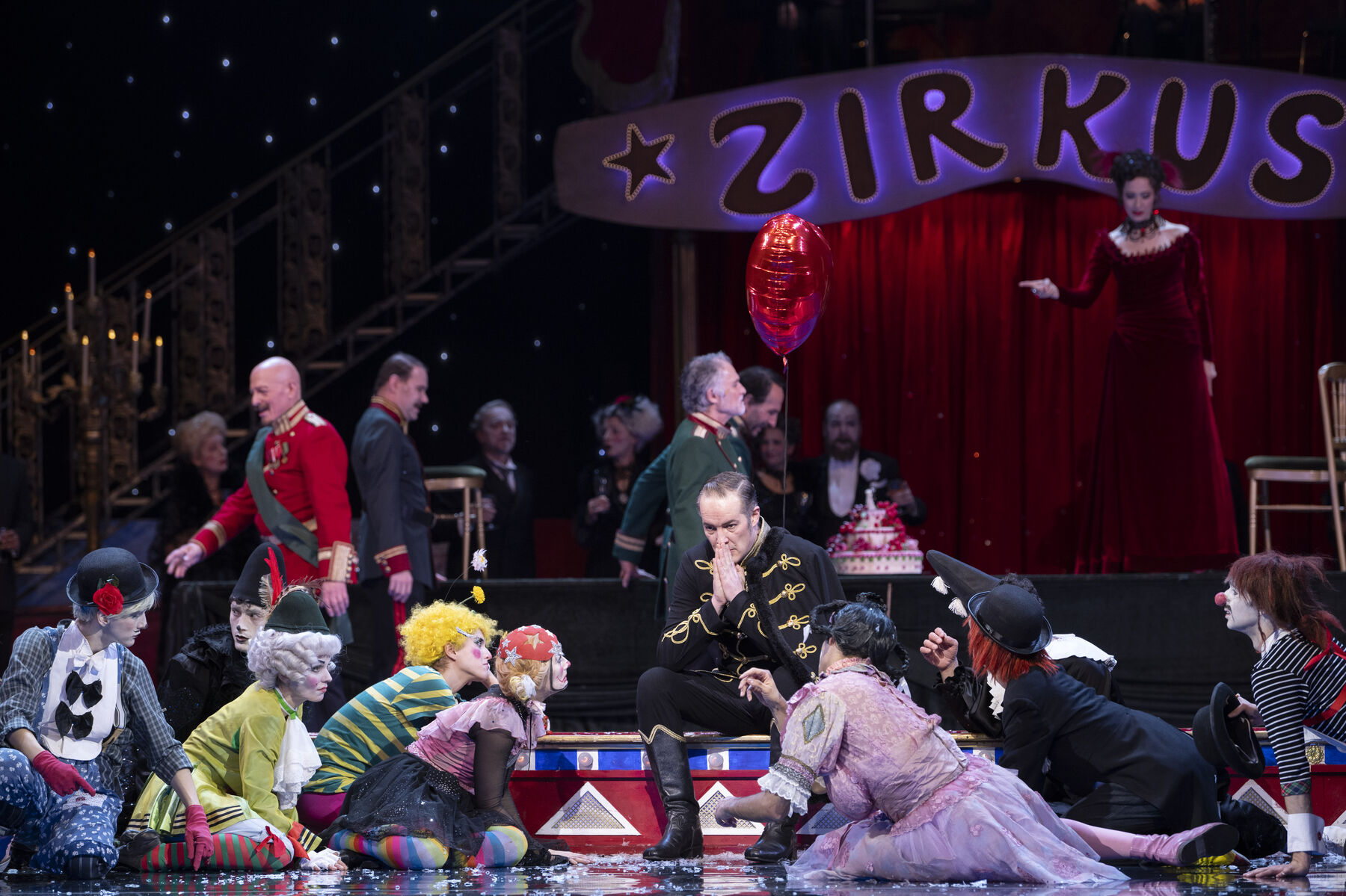 A man sits desperately in front of a full circus ring. In front of him, clowns sit in a semicircle and look at him.