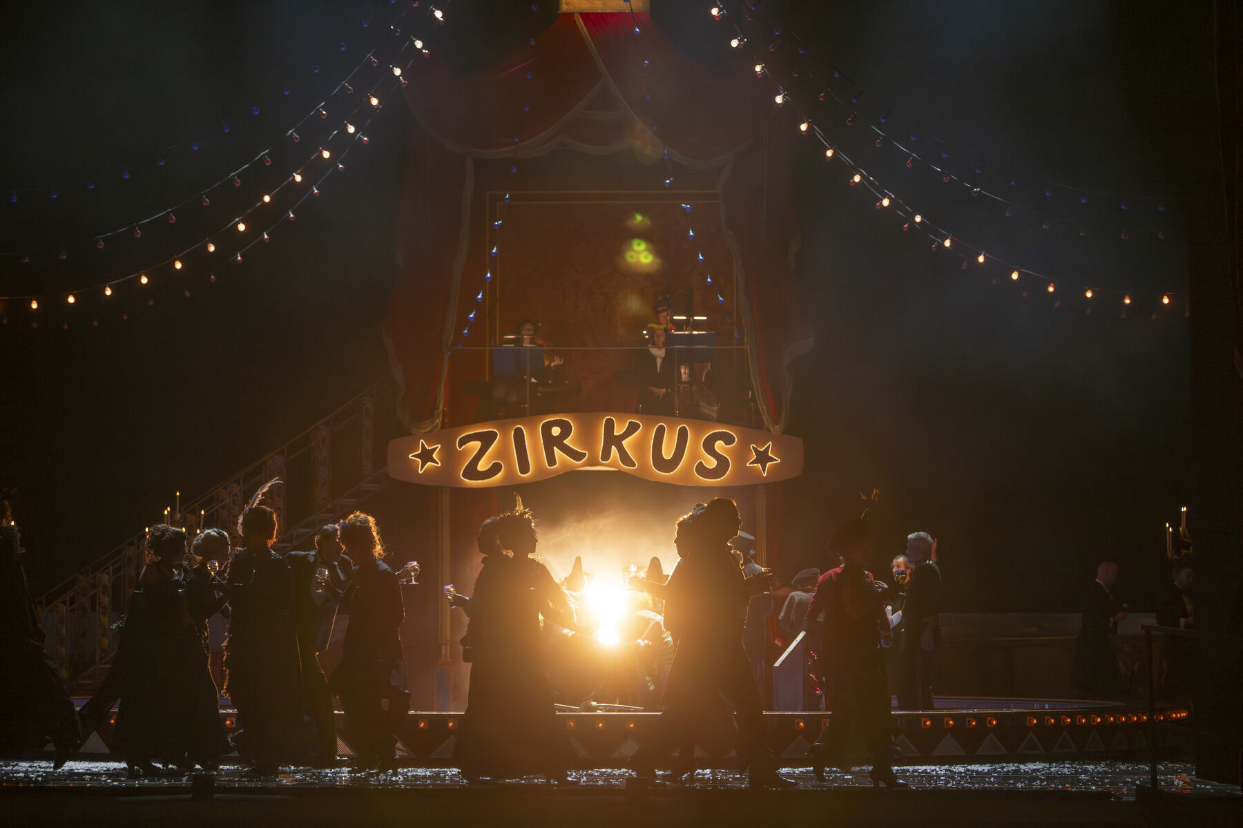A dark circus tent where people dance, illuminated by a single light shining from the background.
