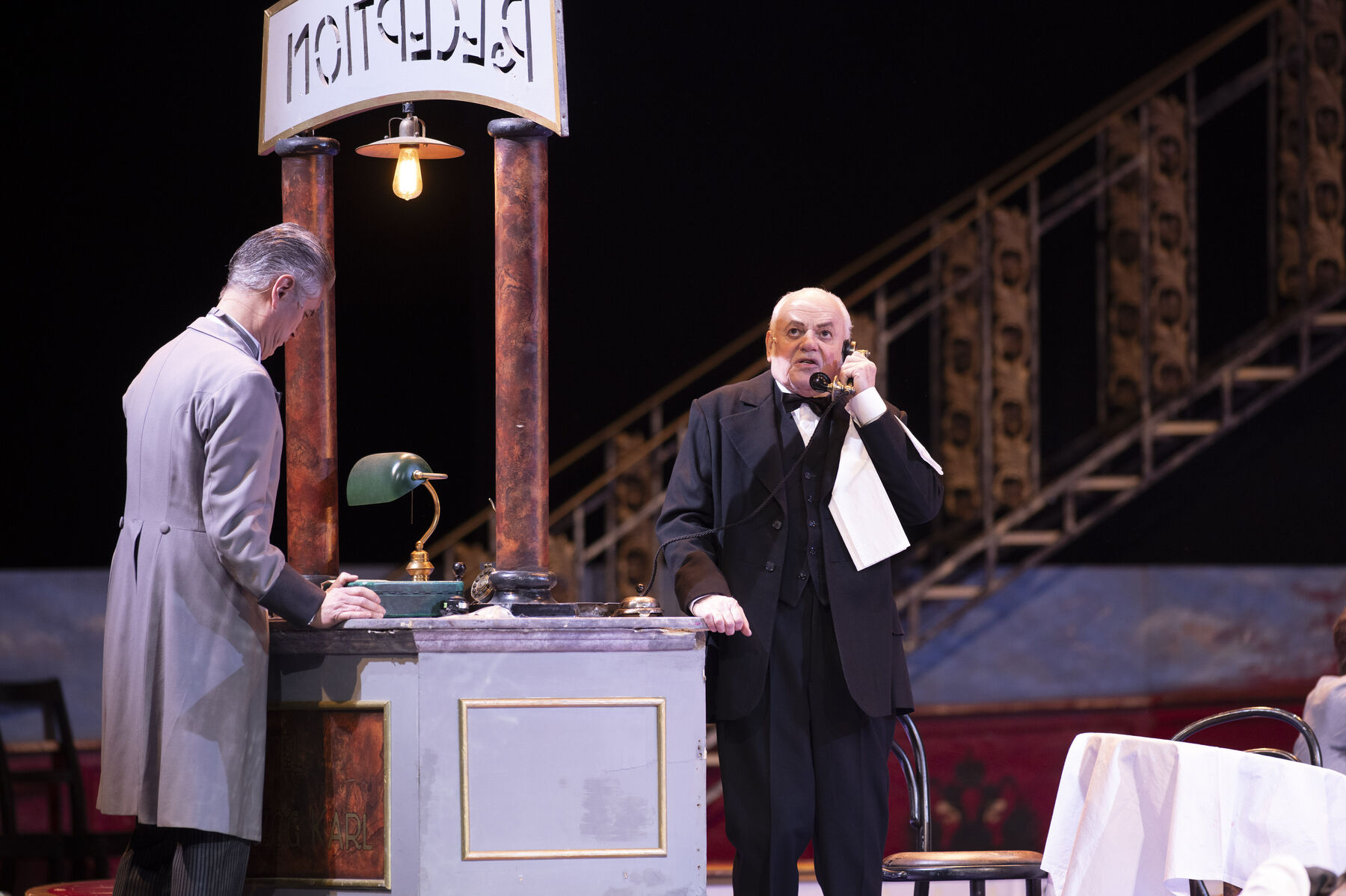 A man in a gray suit is standing at a reception desk. On the other side is a man in a black suit talking on the phone.