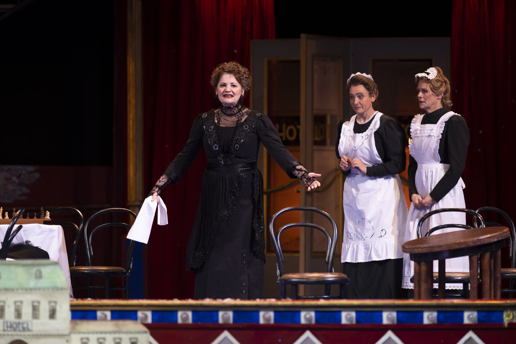 A woman in a black evening gown walks desperately past chairs. Behind her stand two maids in black dresses with white aprons.