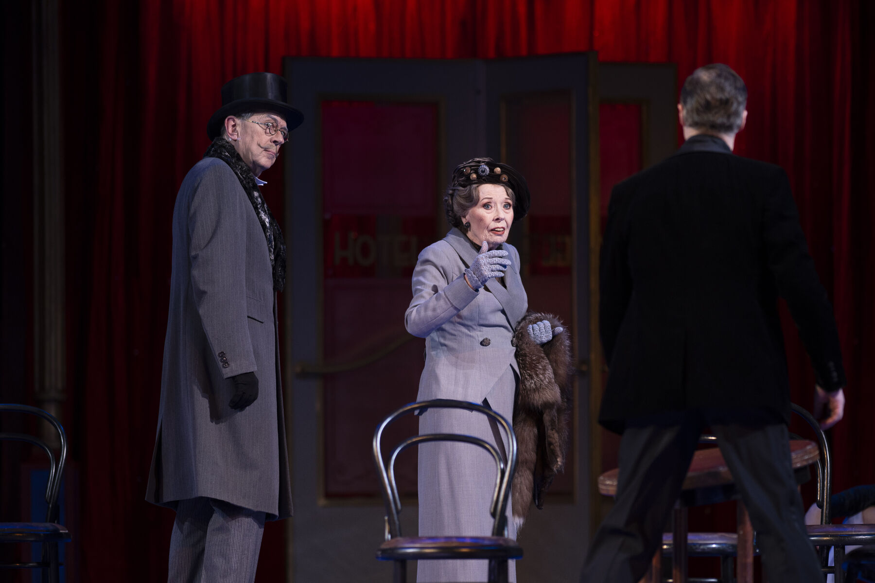 A man and a woman in gray coats are standing behind a chair in front of which a man in a black suit is standing.