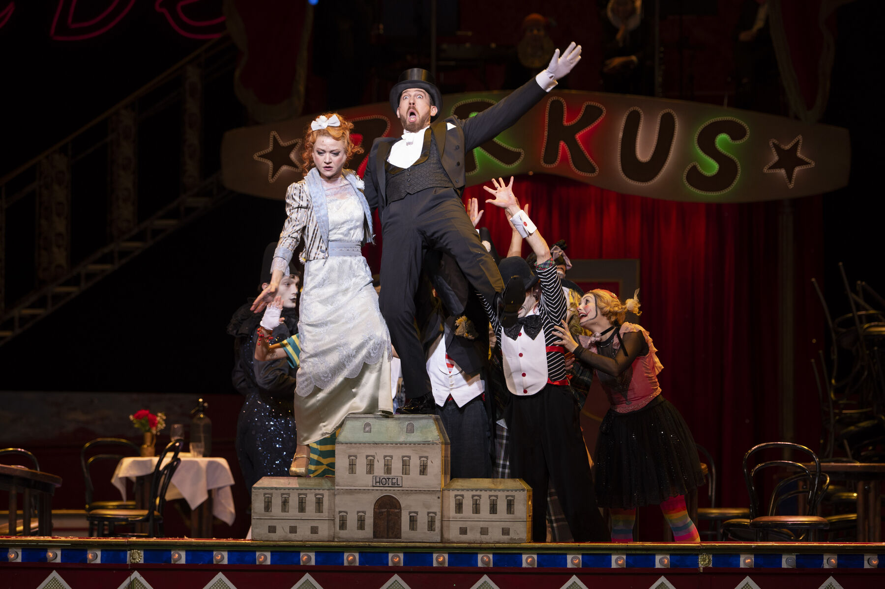 A man in a black suit and black top hat and a woman in a light blue dress are standing on a miniature hotel. Behind them are clowns supporting them.