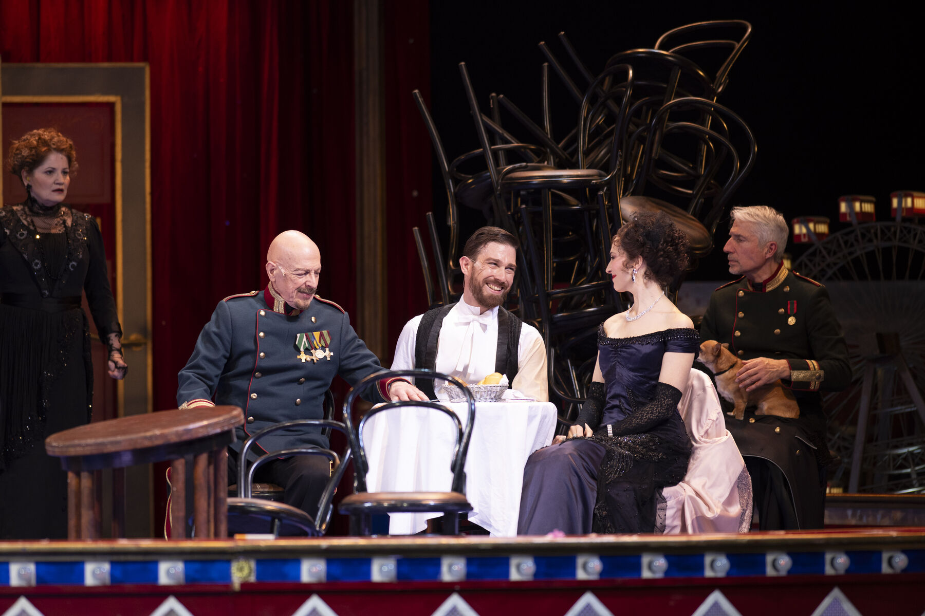 Three elegantly dressed men and a woman in an evening gown are seated around a small, set table.