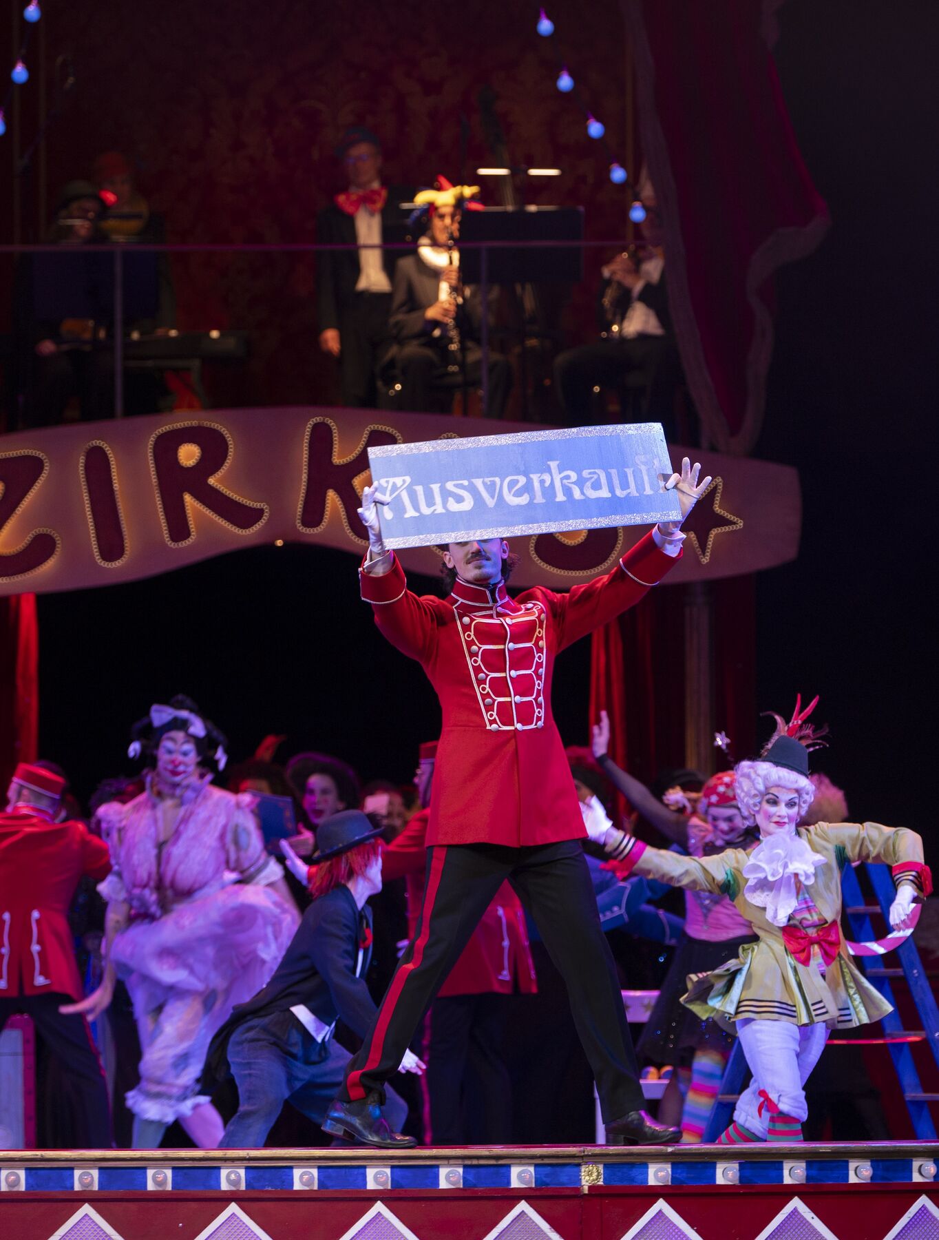 A man in a red uniform stands at the edge of a circus ring. He is holding up a sign that says “Sold Out.” In the background, clowns are dancing.