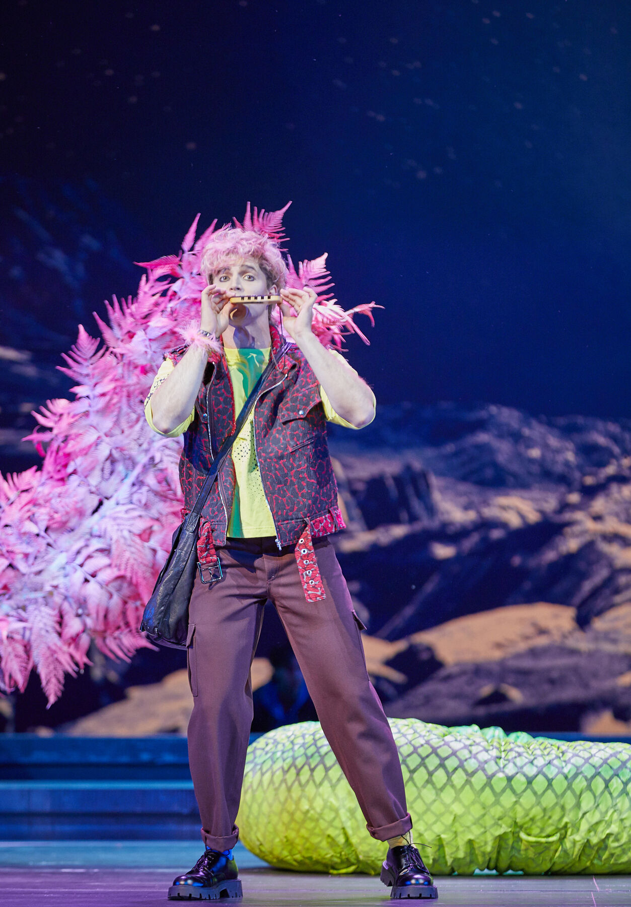 Papageno spielt Panflöte. Er ist bunt gekleidet. Der Mann hat rosa Haare, ein neongrünes T-Shirt, eine dunkelrote Hose und eine schwarz-rot gemusterte Weste an. Im Hintergrund liegt die tote Schlange und ein großer rosa Federbüschel ragt in das Bild. 