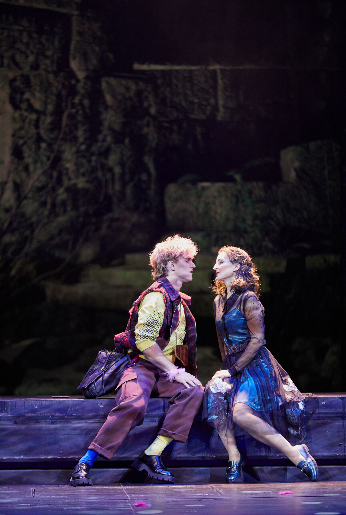 Papageno und Pamina sitzen dicht nebeneinander. Sie lehnen sich beide leicht nach vorne und nähern sich einem Kuss an. Im Hintergrund sieht man eine Steinwand.