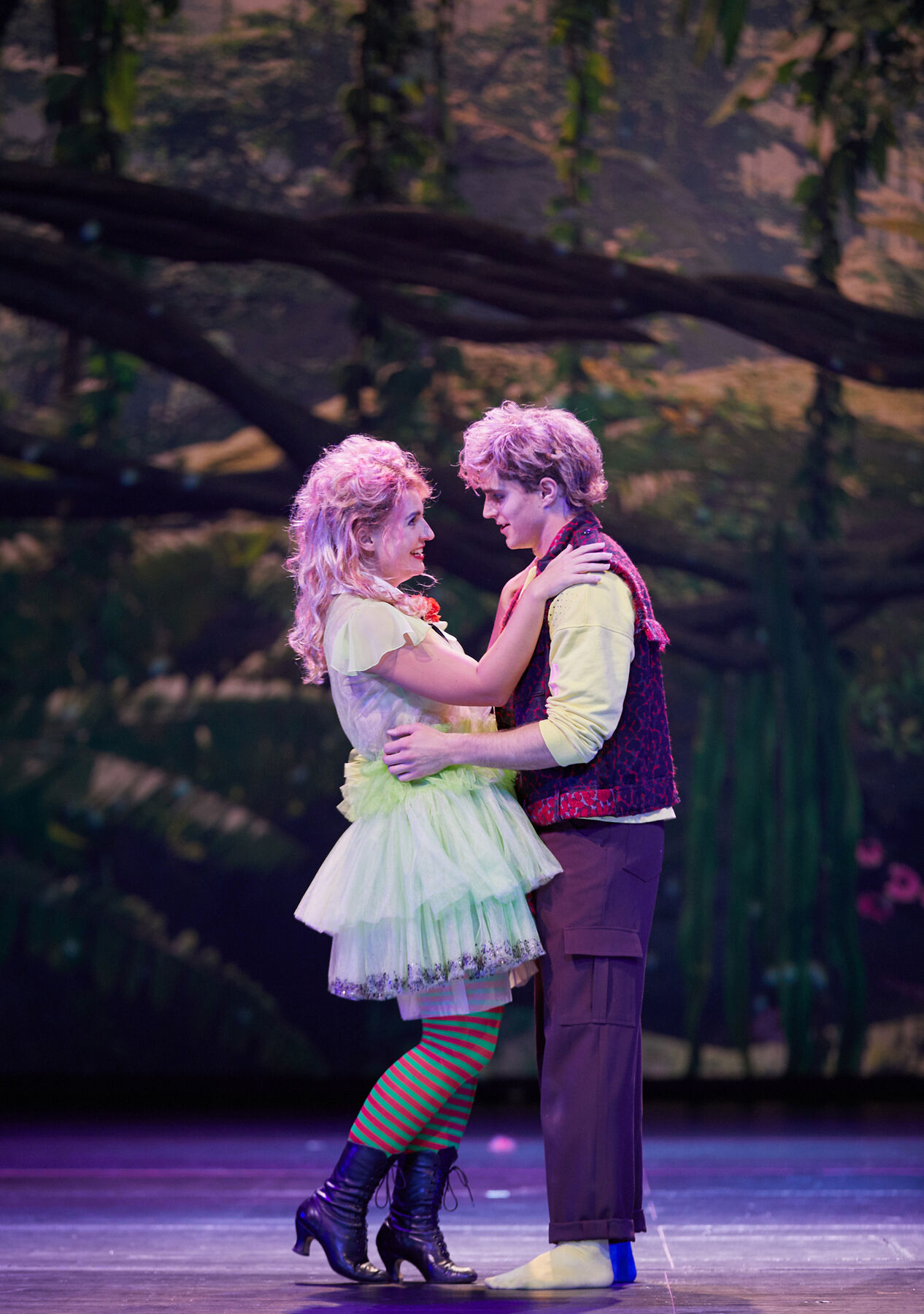 Papagena und Papageno stehen sich glücklich gegenüber. Sie legt ihre Hände auf seine Schultern und er seine um ihre Taille. Das Licht ist leicht rosa. Im Hintergrund sieht man von Ranken umwobene Äste. 