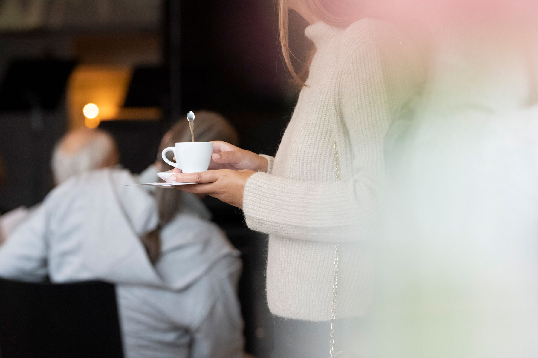 Eine Frau hält einen weißen Unterteller mit einer weißen Kaffeetasse aus der ein silberner Löffel ragt. Sie trägt einen beigen Pullover. Im Hintergrund sitzen zwei Personen.