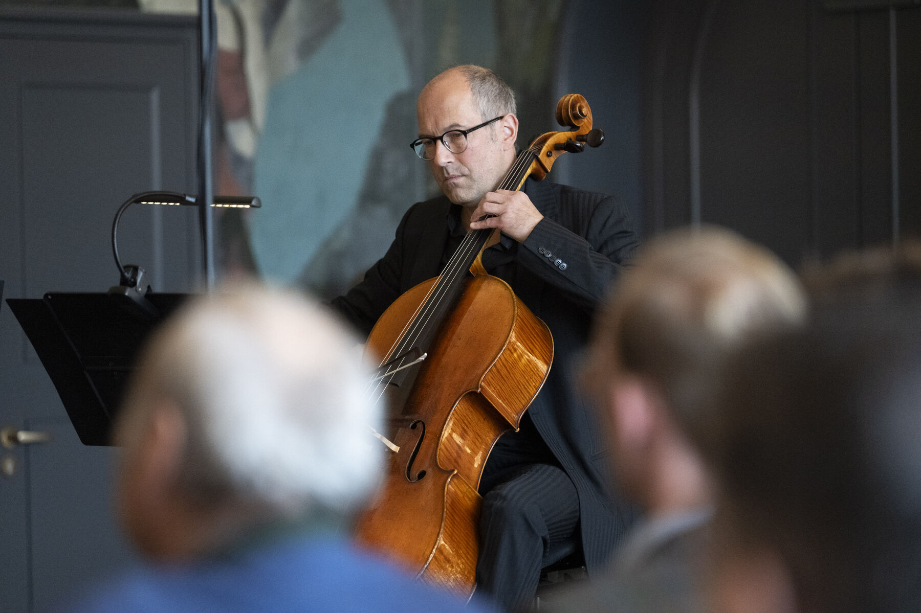 Unscharf im Vordergrund sitzen zwei Zuschauer, die auf die Bühne des Pausenfoyers des Gärtnerplatztheaters schauen. Scharf im Hintergrund sind sitzt ein Mann des Orchesters des Staatstheaters am Gärtnerplatz und spielt auf einem Cello.