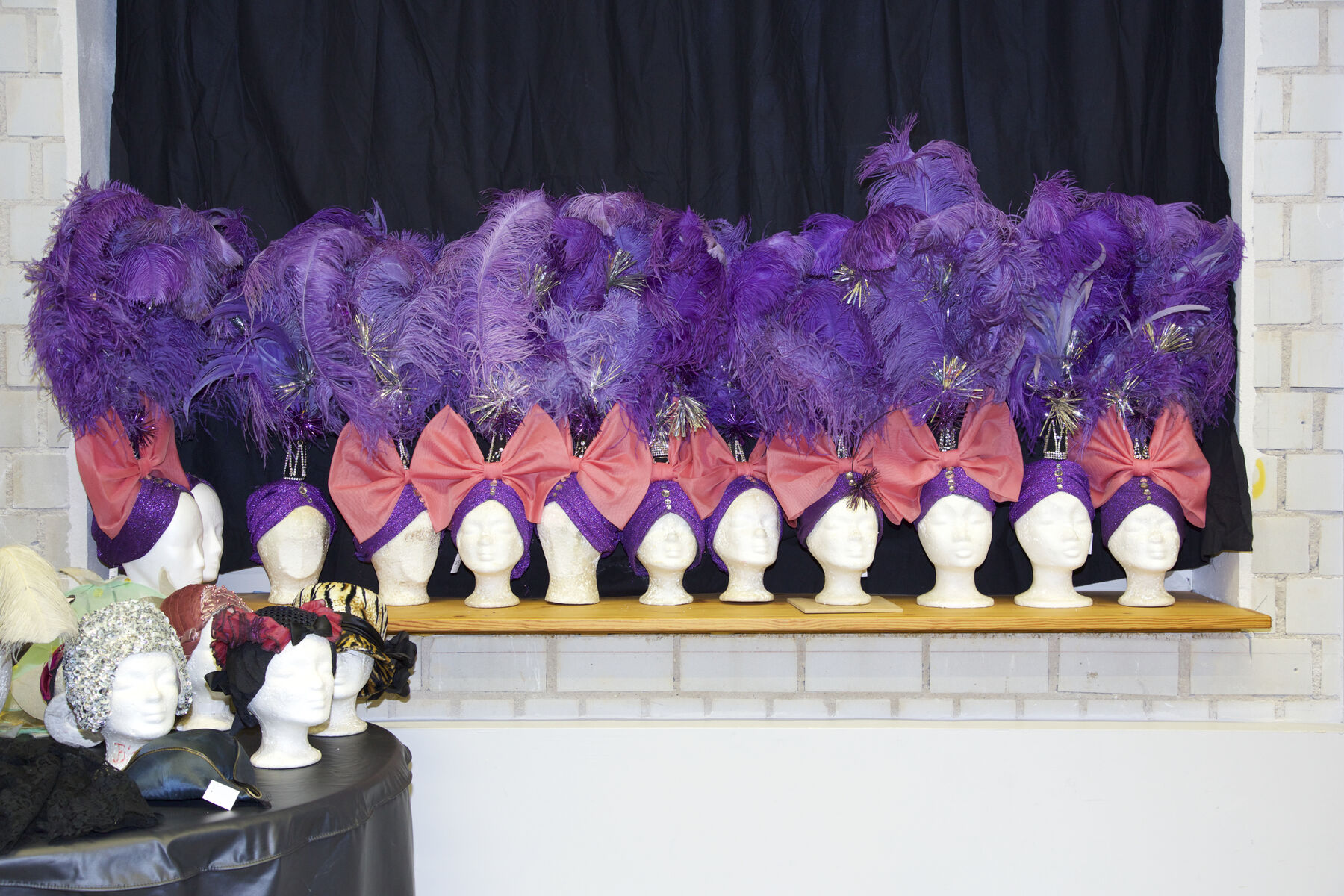 Auf Modellköpfen sind extravagante Kopfbedeckungen ausgestellt. Die Kopfbedeckungen sind violett und darauf sind große, pinke Schleifen positioniert. Darüber hinaus, sind violette Federn Teil der Kopfbedeckungen. 