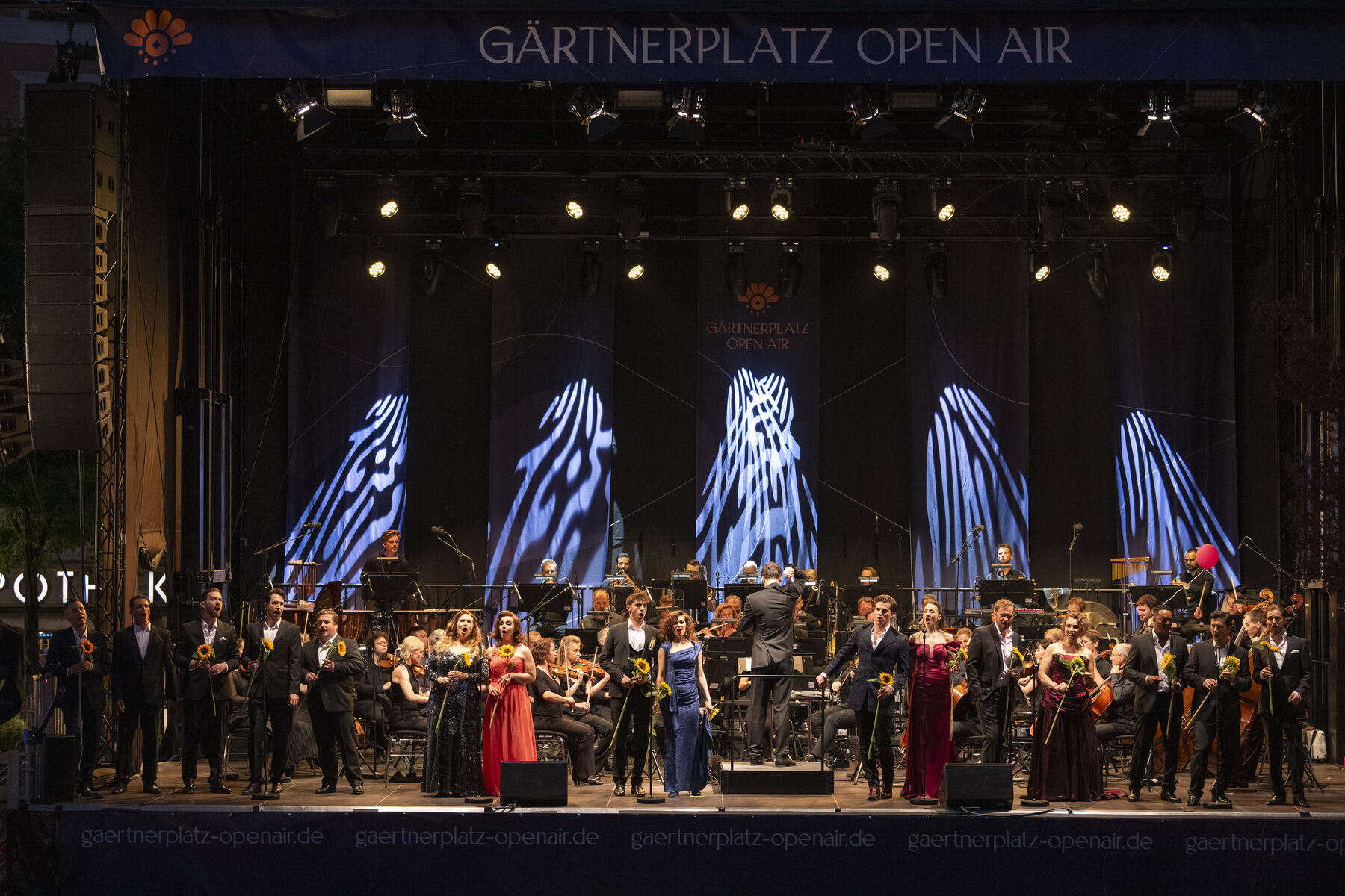 A group of singers, an orchestra, and a conductor stand on an open-air stage in front of the Gärtnerplatztheater.