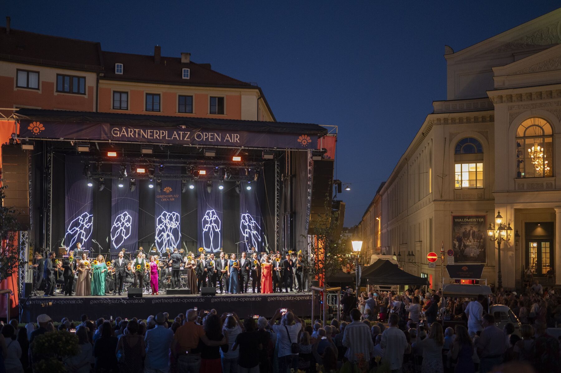 Sänger und Sängerinnen stehen in Abendgarderobe auf einer Freilichtbühne. Hinter ihnen ist ein Dirigent und ein Orchester zu sehen.