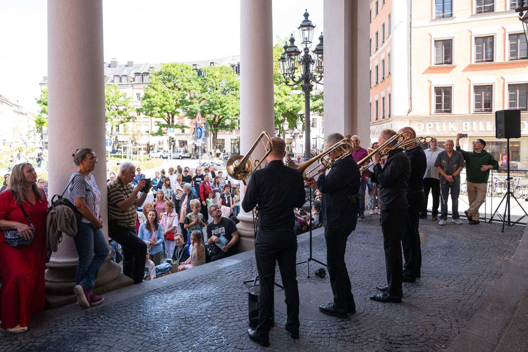 Vier Posaunisten stehen vor dem Gärtnerplatztheater und spielen eine Fanfare. Vor ihnen stehen viele Menschen und hören zu.