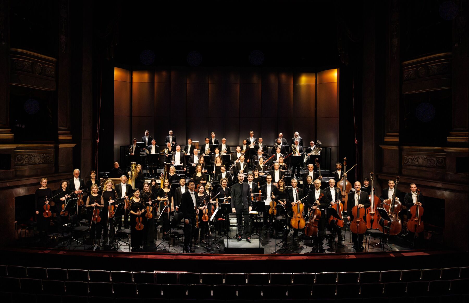 Das Orchester des Staatstheaters am Gärtnerplatz steht  mit ihrem Dirigenten auf einer hell erleuchteten Bühne, die Instrumente in der Hand und lächelt in die Kamera.