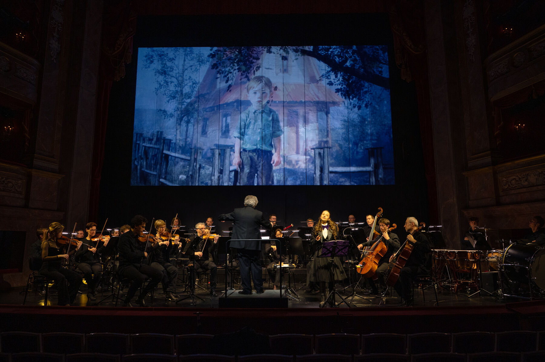 Auf der Bühne befinden sich ein Orchester, der Dirigent und die Moderatorin Marina Blanke. Über ihnen ist auf der Leinwand Peter zu sehen, wie er das Gartentor offen gelassen hat.