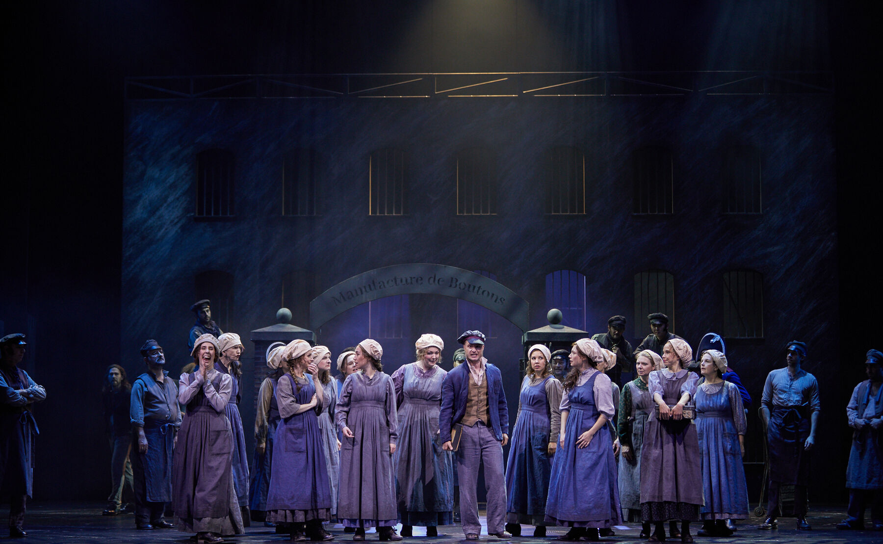 Eine Gruppe von lila und blau bekleideten Frauen stehen um einen Mann, der in lila Hose und beiger Weste mit blauer Jacke nach vorne blickt und singt. Die Frauen schauen sich an und reden miteinander. Im Hintergrund erkennt man die Silhouette eines Gefängnisses und dessen Eingangstor.